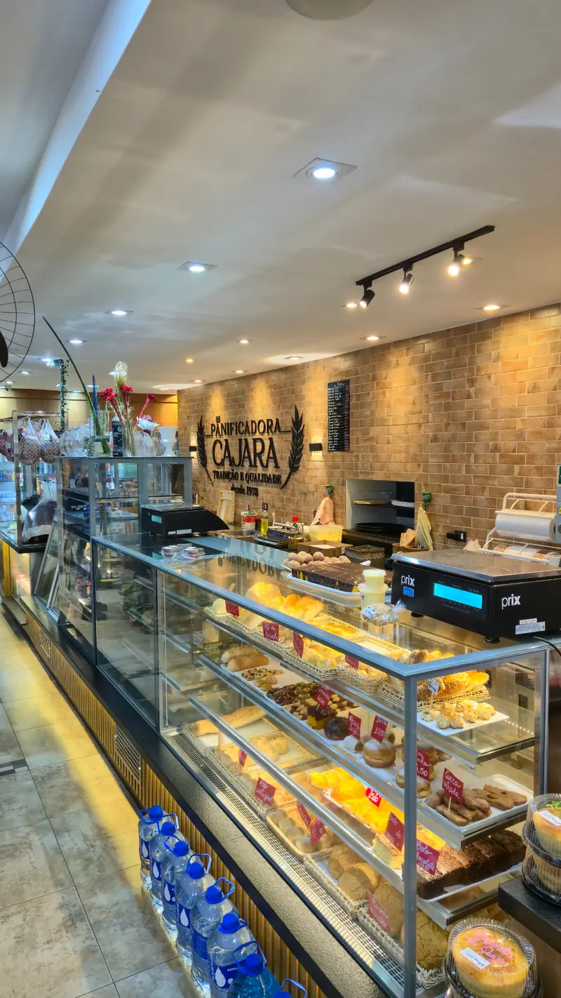 Vitrine iluminada da Panificadora Cajara com pães, doces e o logotipo na parede de tijolinhos