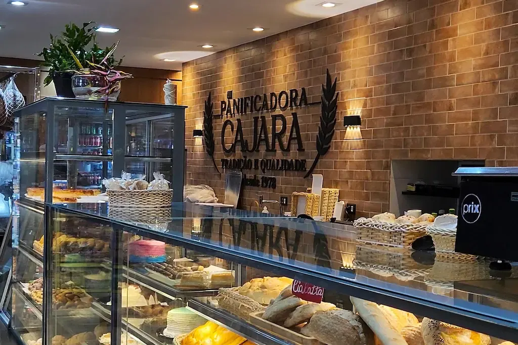 Vitrine de bolos e doces em frente à parede de tijolinhos com o logotipo da Cajara